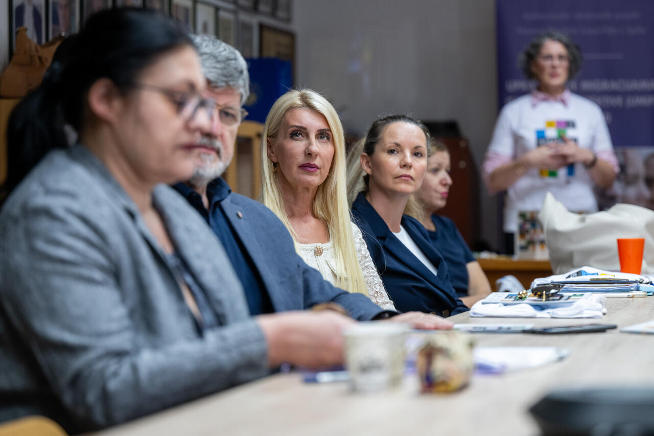 Project members seated at the table while a colleague presents in the background.