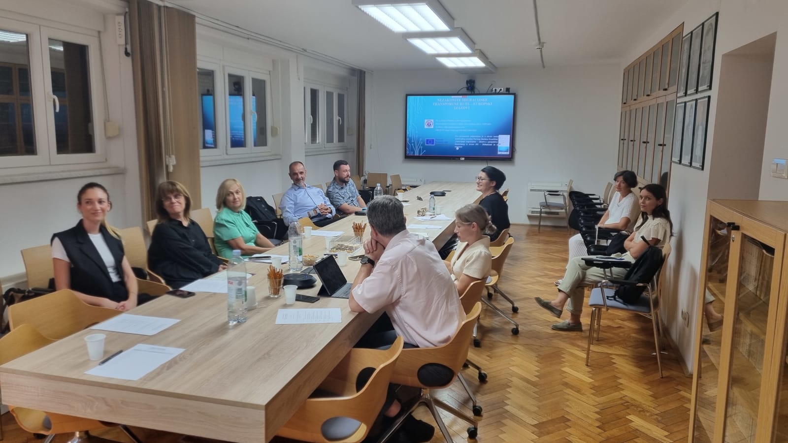 Participants seated around the table during a discussion, with a presentation on illegal migration routes on the screen.